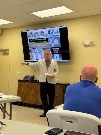 NSPIRE Training. The back of a man seated in a chair while Dennis DiBello is standing in front of a smartboard with the training on it.