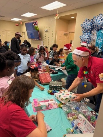 Children standing around tables with presents and adults helping with presents. 