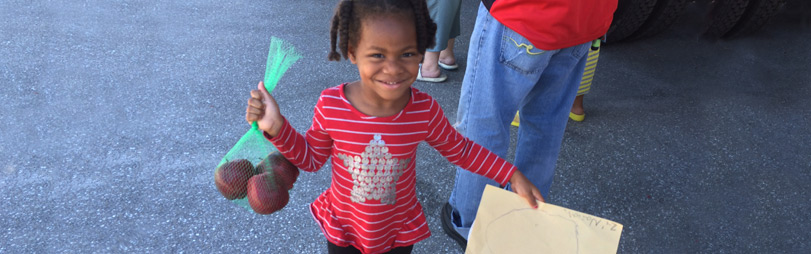 A little girl holding a bag of 3 red apples in her right hand and a portrait in her left hand.