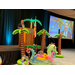  A man standing behind a podium on a stage with palm tree inflatables and other floaties around it. 